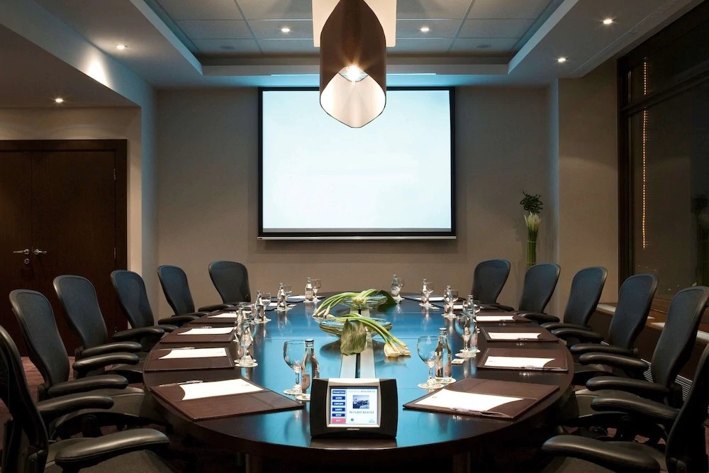 An image of a meeting room with chairs around a oval table and large screen on the front wall.